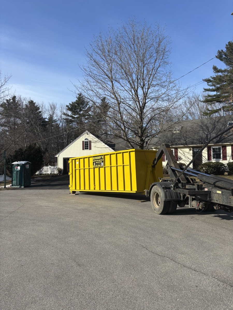 Yellow Scribby's 15-yard dumpster being set on a paved driveway in front of a small white colonial with brick chimney in southern NH.