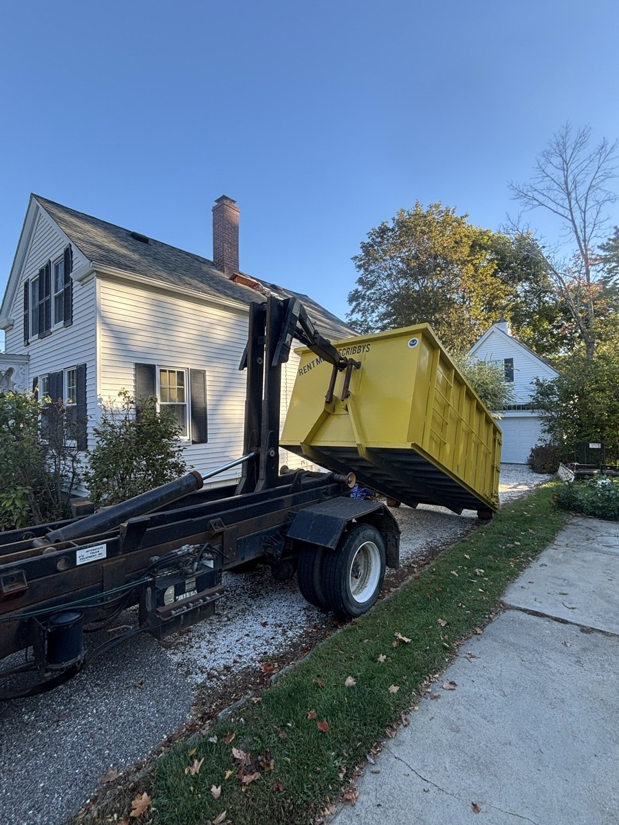 Yellow Scribby's 20-yard roll-off dumpster being placed on the driveway of a white New England Cape with brick chimney during early fall foliage.