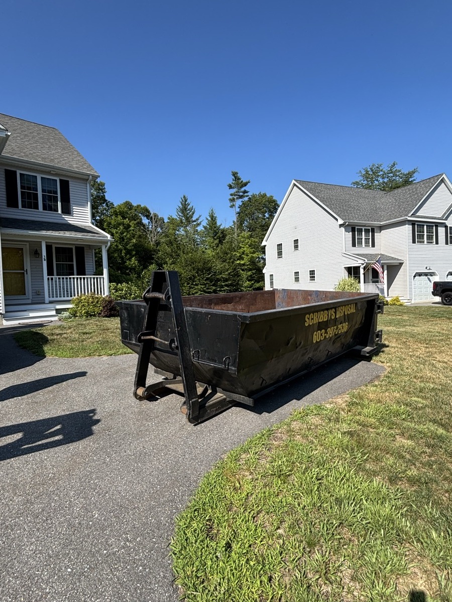 Scribby's 20-yard roll-off dumpster being set on the asphalt driveway of a white colonial home with American flag in southern NH.