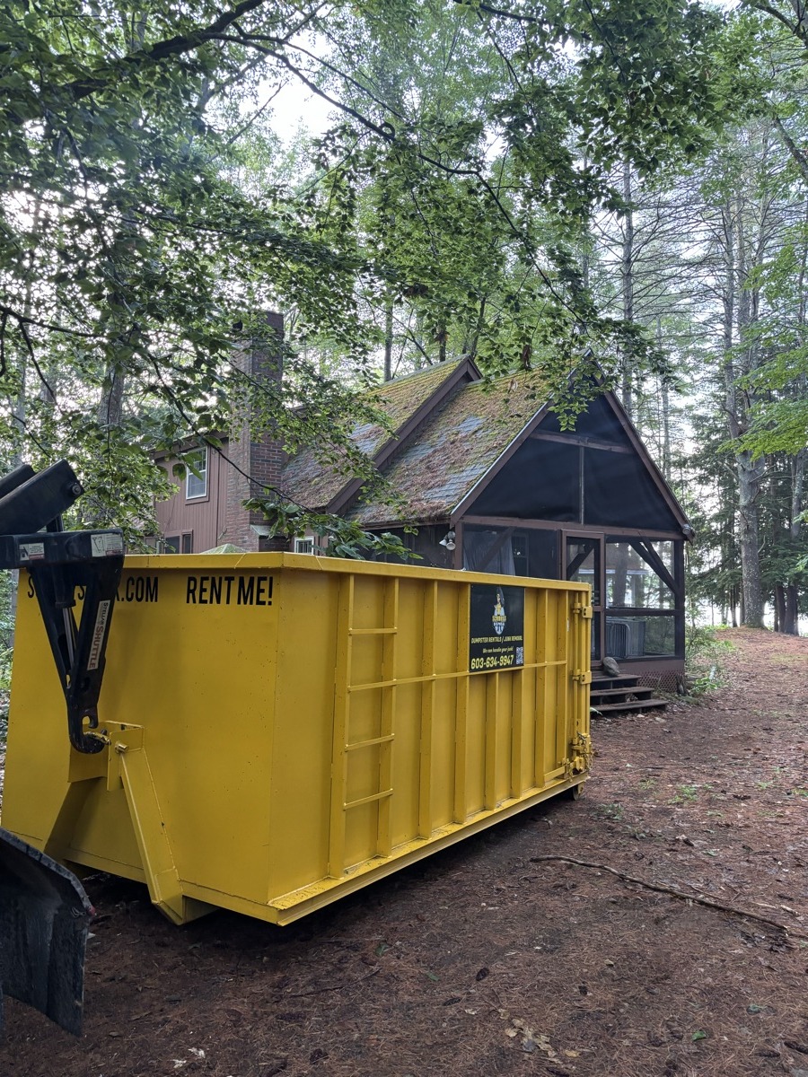 Yellow Scribby's 'Rent Me' dumpster set on dirt in a dense southern NH woods next to a small cabin — rural teardown cleanup.