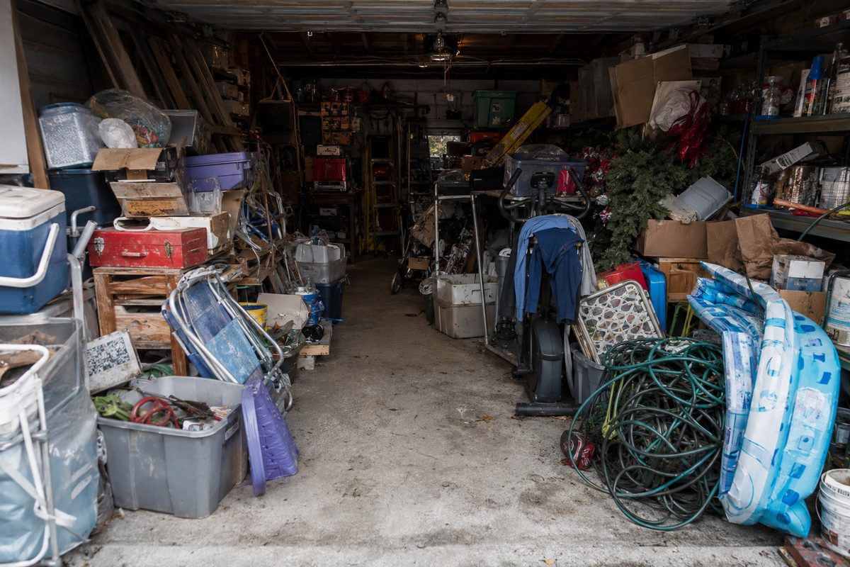 Cluttered garage interior with bins, paint cans, exercise bike, and holiday decorations packed floor to ceiling along a narrow path.