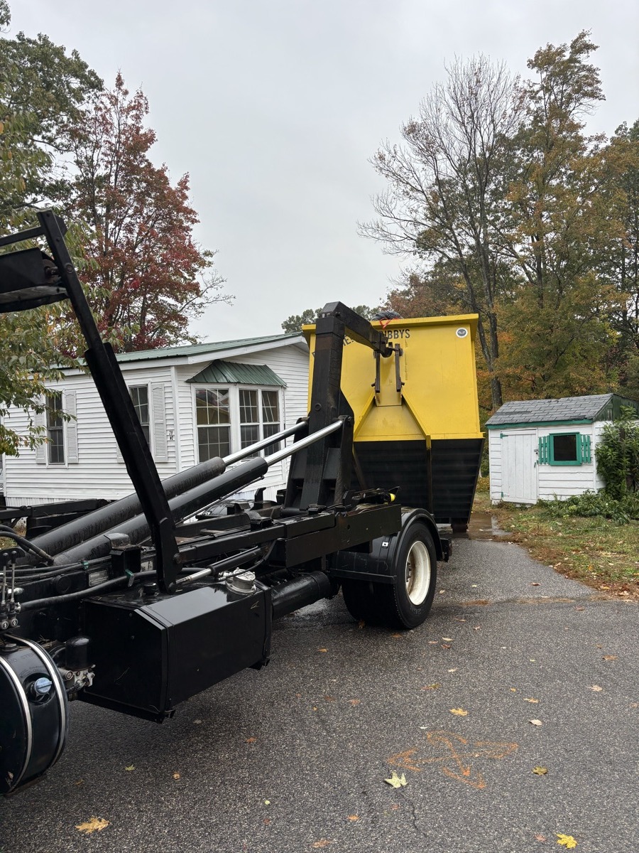 Scribby's roll-off dumpster being delivered in front of a small white antique farmhouse with fall foliage in rural southern NH.