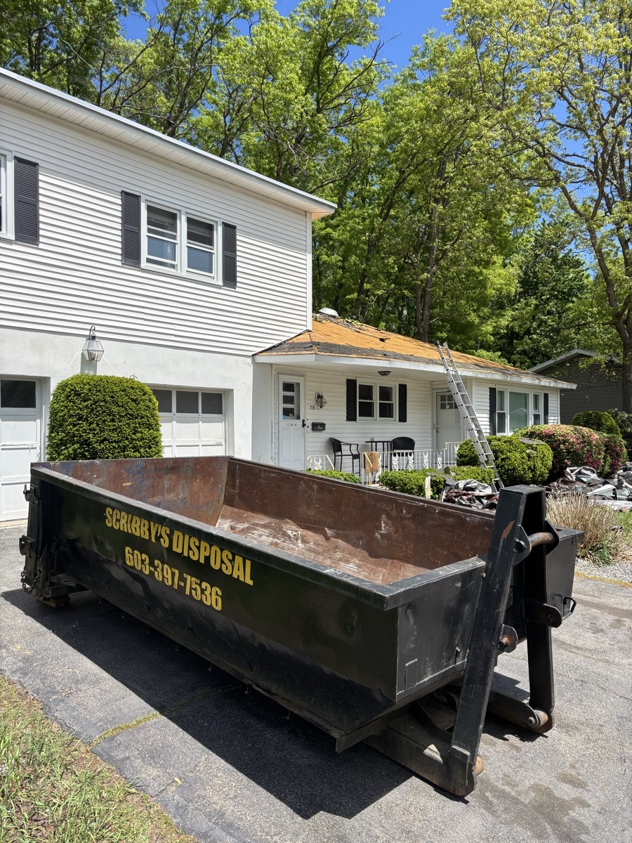 Black Scribby's Disposal dumpster being placed by truck in front of a long white colonial with manicured boxwood shrubs — upscale southern NH estate.