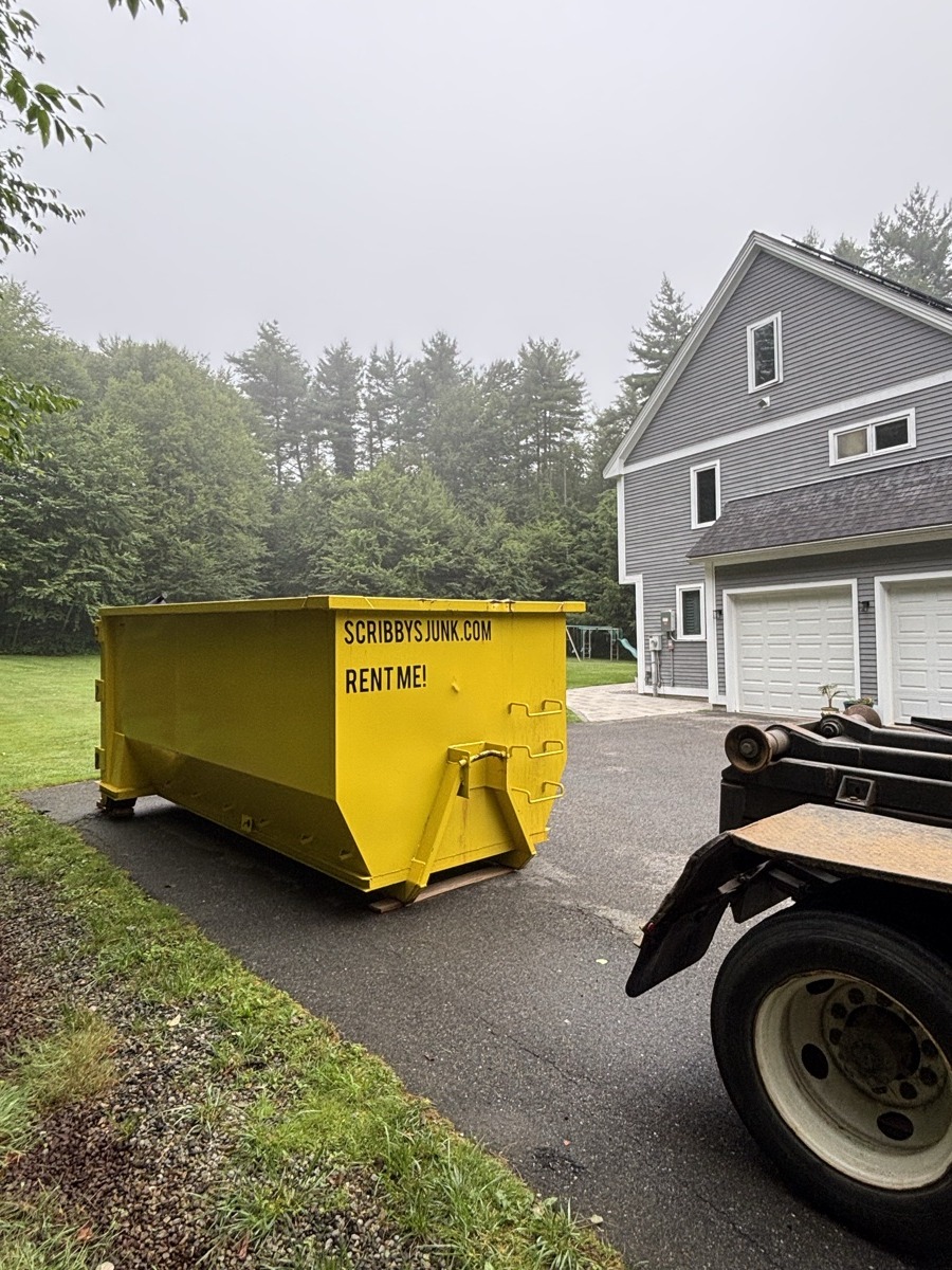 Scribby's roll-off dumpster being placed in front of a gray-vinyl ranch home with a two-car garage on an overcast NH day.
