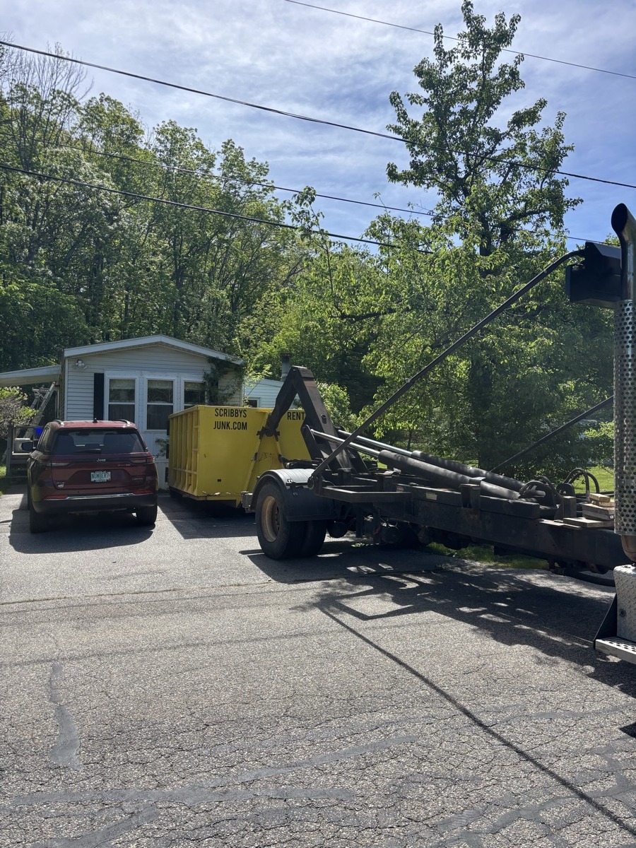 Yellow Scribby's Junk dumpster being placed by truck in front of a small white house with a red sedan parked in the spring driveway.