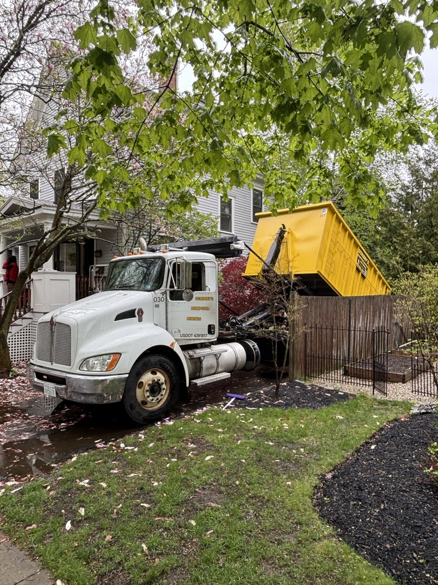 Scribby's roll-off truck delivering a yellow dumpster to a mulched residential driveway in southern NH on a leafy spring day.