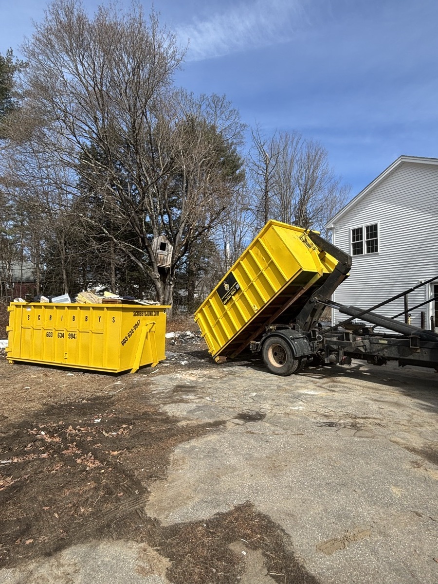 Two yellow Scribby's dumpsters at a sandy southern NH yard — one mid-placement off the truck, showing the range of available sizes.