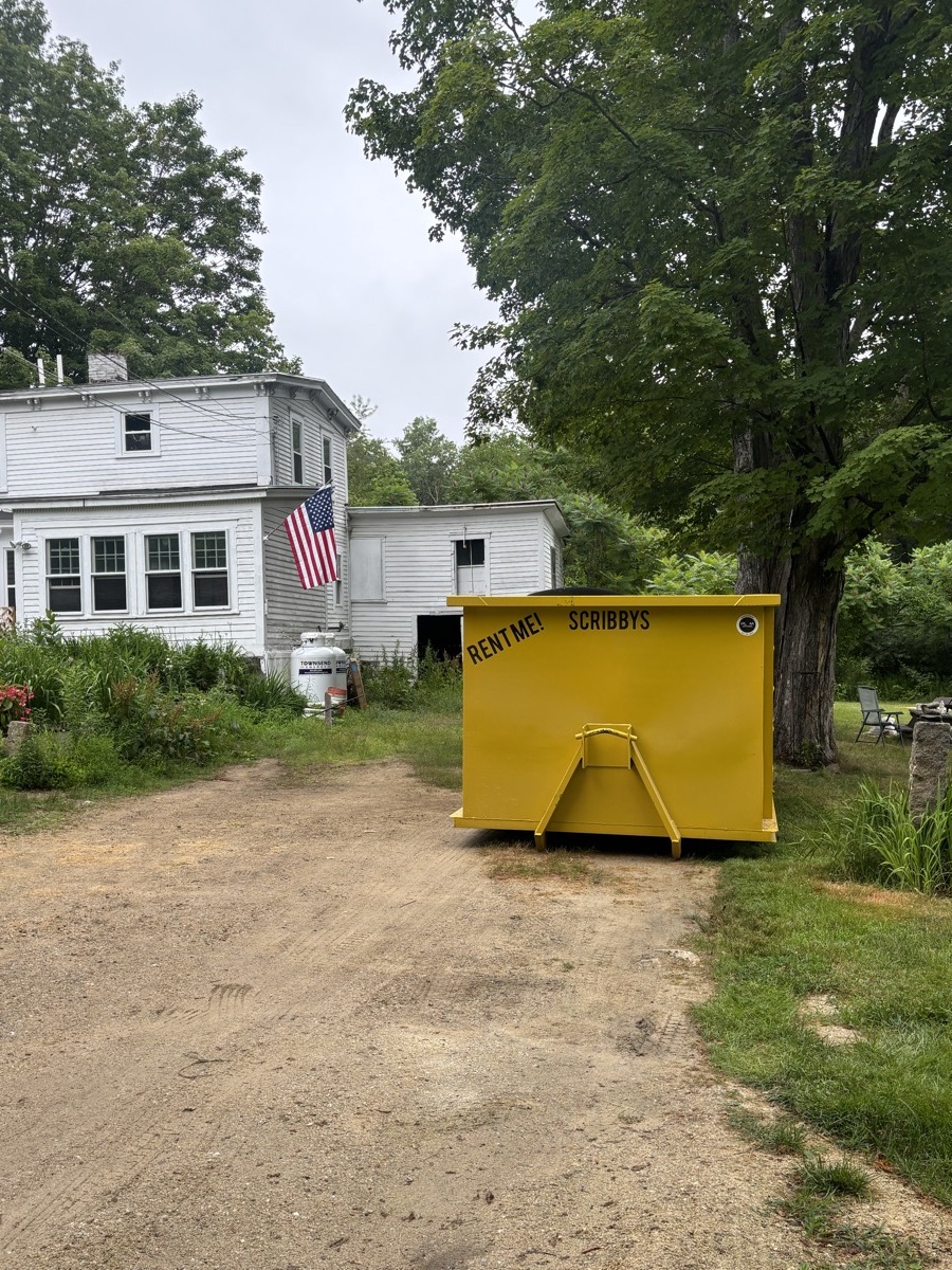 Yellow Scribby's 'Rent Me' dumpster on a dirt and gravel pad in front of a white antique farmhouse in rural southern NH.
