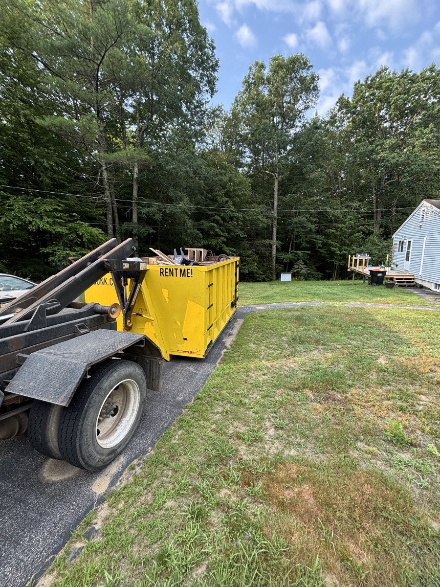 Yellow Scribby's 'Rent Me' dumpster on a gravel driveway at a modest rural southern NH home with grass and trees.