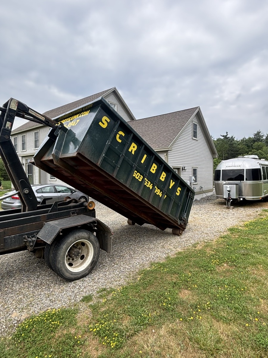 Scribby's hook-lift truck setting a roll-off dumpster on a gravel driveway at a rural modern-barn-style home in southern New Hampshire.