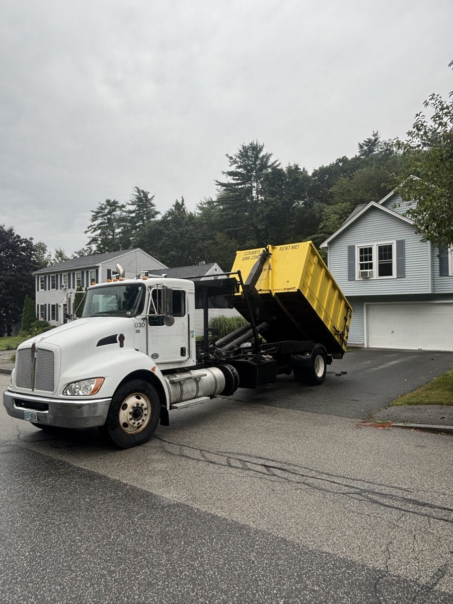 Scribby's roll-off truck delivering a yellow dumpster to a small gray ranch home on an overcast day in southern NH.