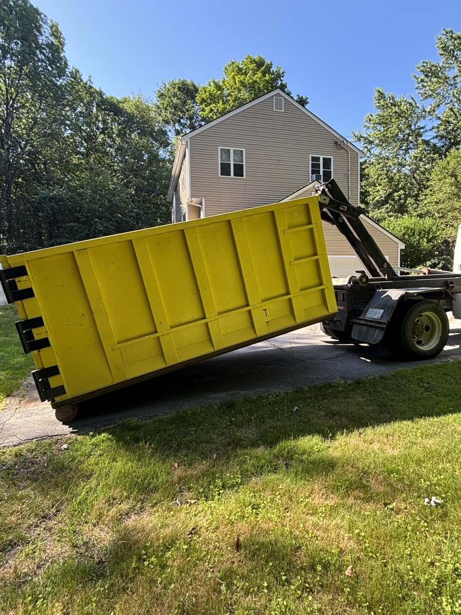 Yellow Scribby's roll-off dumpster being placed on a driveway next to a grassy field with a beige barn-style building in southern NH.
