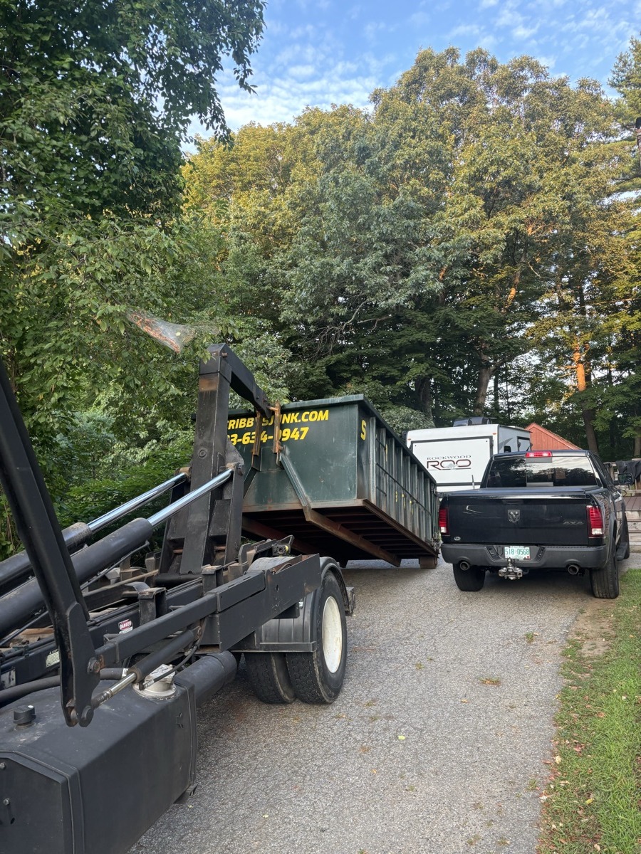 Scribby's roll-off dumpster being set on a suburban driveway with tall NH pines in the background.