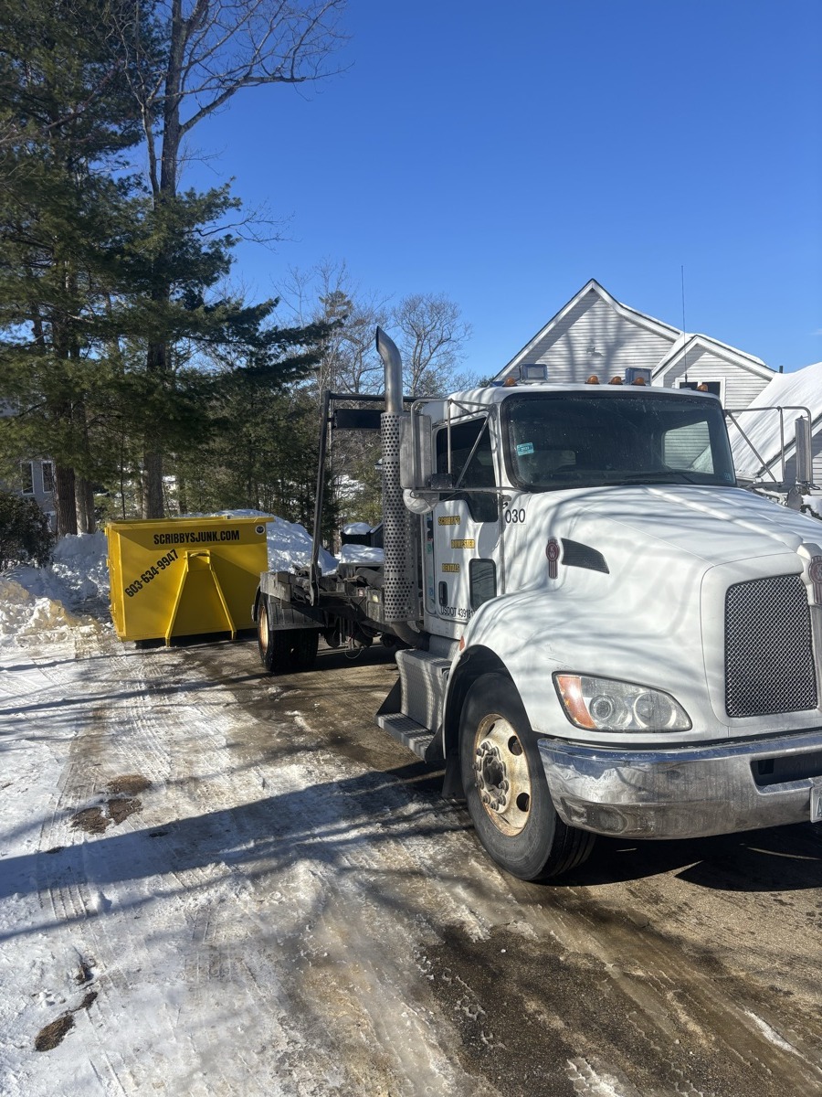 Front three-quarter view of Scribby's white roll-off truck with yellow dumpster on the bed, parked on a sunny southern NH driveway.