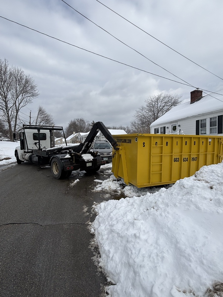 Yellow Scribby's roll-off dumpster being placed on a snowy driveway with tall snowbanks in front of a white southern NH home.