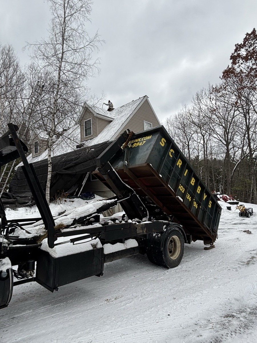 Black Scribby's roll-off with snow on top, mounted on a white Scribby's truck, parked beside a small southern NH home in heavy snow.
